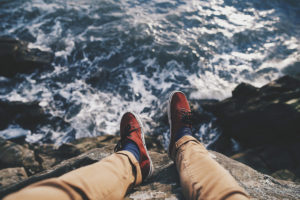 A person's legs dangling over a rocky ledge, with the ocean waves crashing below.