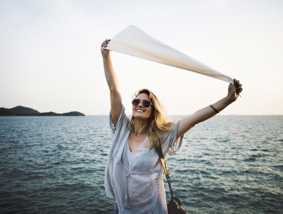 A joyful woman holding a white scarf above her head, smiling with the ocean in the background on a sunny day.